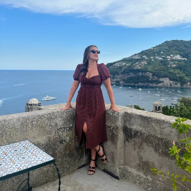 Travel Advisor Carissa Bartoli with a dark red dress in front of a ledge with water and green lush hills in the background.