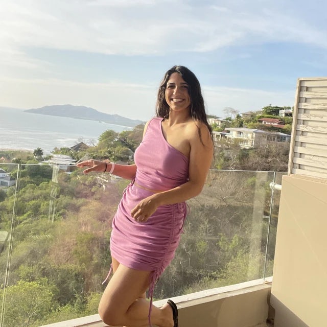 Fora travel agent Tali Gonzalez wearing pink dress standing on balcony overlooking green hills and ocean