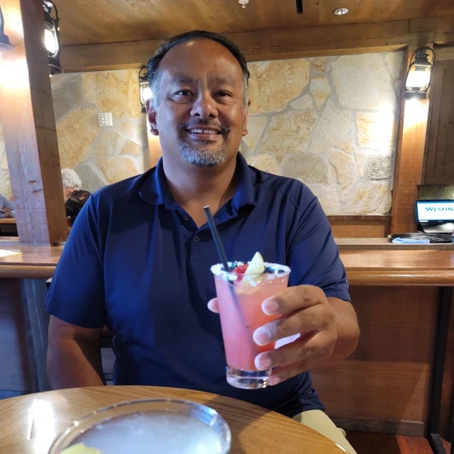 Travel Advisor John Chamberlain holds up a pink cocktail at a bar with a map all the wall behind him