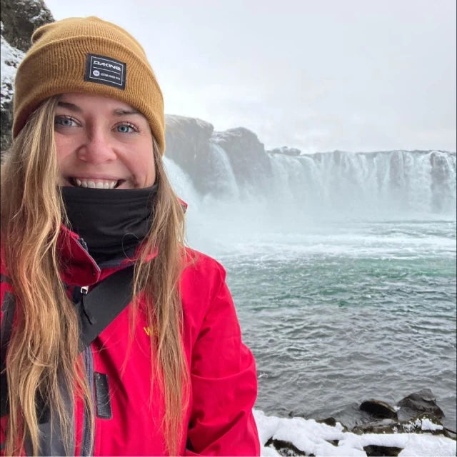Travel Advisor Lindsey Leichthammer stands in front of a waterfall in a snow covered shore wearing a winter hat and red jacket