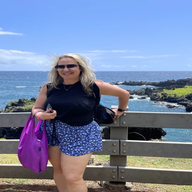 Travel Advisor Lauren Kaskiel stands against a wooden fenceon a cliff over the ocean wearing blue shorts and a black tank top