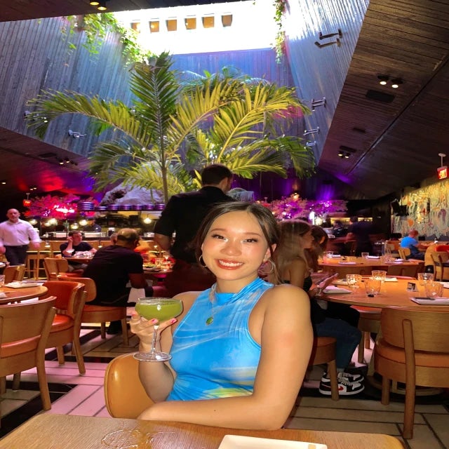 Travel Advisor Tiffany Nguyen wears a blue top and holds up a green cocktail at a restaurant with a large palm tree 