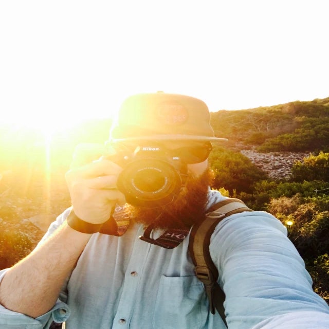 Travel Advisor Jeff Karges holds a camera up to his eye with the sun peaking out behind his shoulder wearing a straw hat 