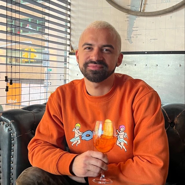 Travel Advisor Omar Gonzalez with an orange sweatshirt and holding an orange drink in front of a map on the wall behind him.
