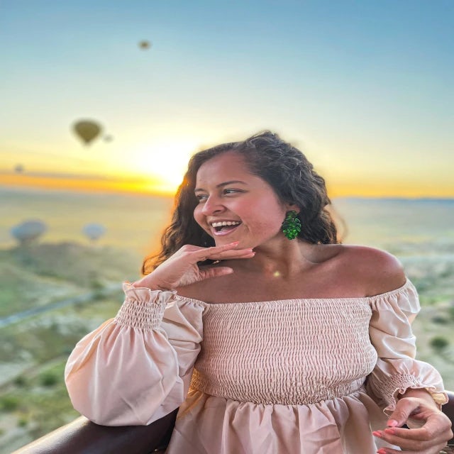 Travel Advisor Rosealety Galea stands on a balcony at sunset wearing a light pink dress with hot air balloons floating through the sky. 