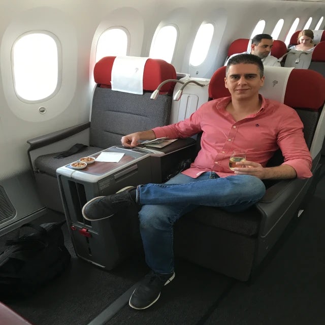 Fora travel agent Boris BK wearing red shirt and jeans sits in airplane seat