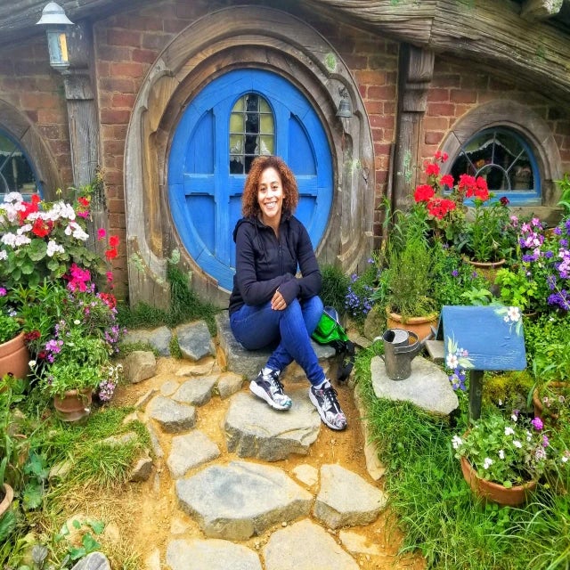 Carly Turner Travel Agent wearing blue jeans and black jacket sitting in front of brick house with blue, circular door