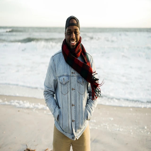 travel advisor Krystofer Henry in a light denim jacket and red scarf smiling on an ocean beach