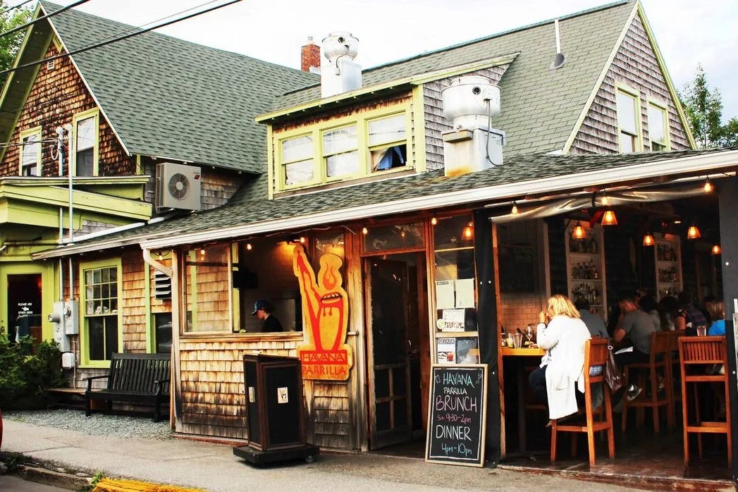 The exterior of a restaurant with wood paneling, yellow trim and people dining on counter height stools next to a chalkboard sign. 