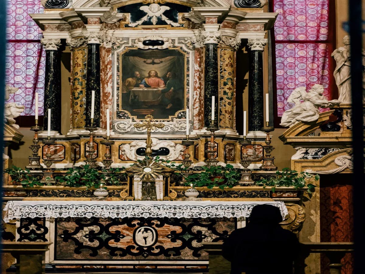 A person in a black robe standing in front of a gold and black altar in a cathedral.