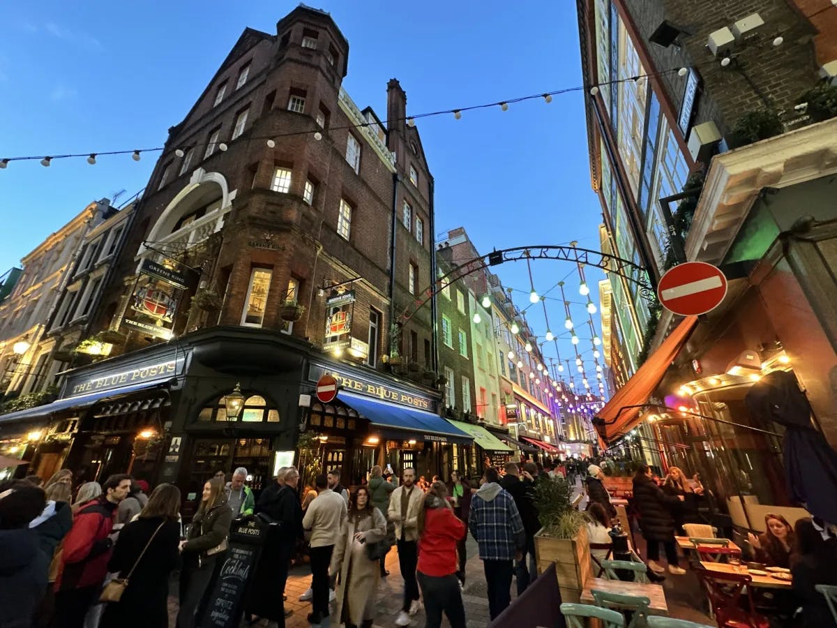 An image of an outdoor shopping center in London with various buildings, string lights, moving lights, people shopping and signage with awnings in the evening. 