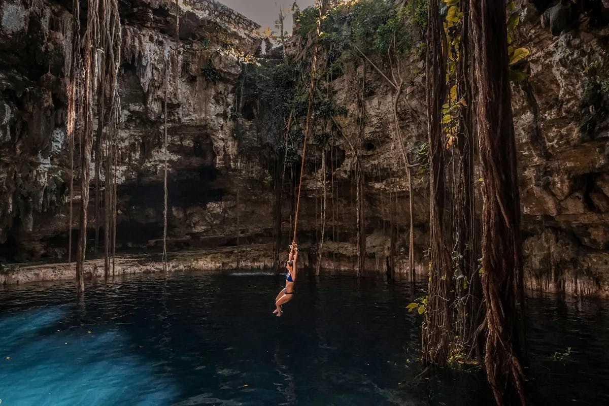 A woman in a blue bathing suit swinging from a rope into a cenote in Maya Riviera Mexico.
