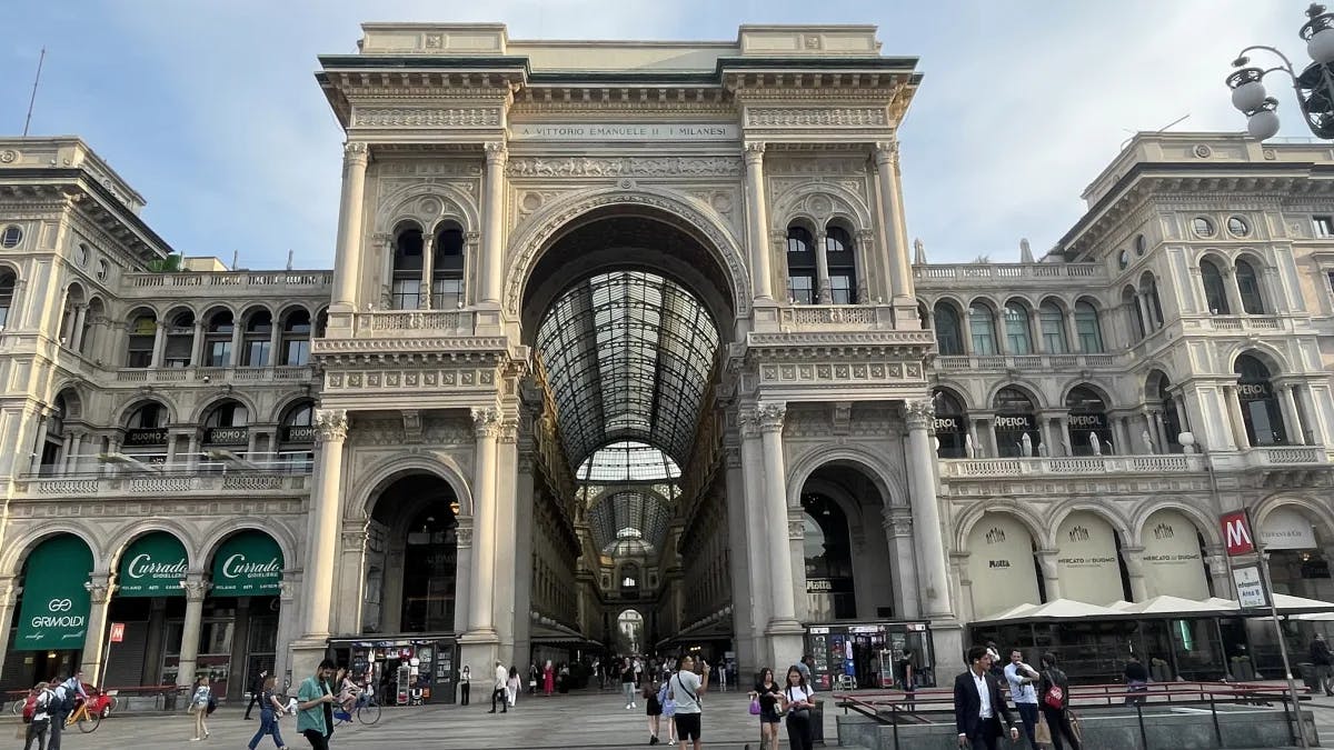 A view of the large, stone shopping center in Milan with people walking around outside in front of it. 