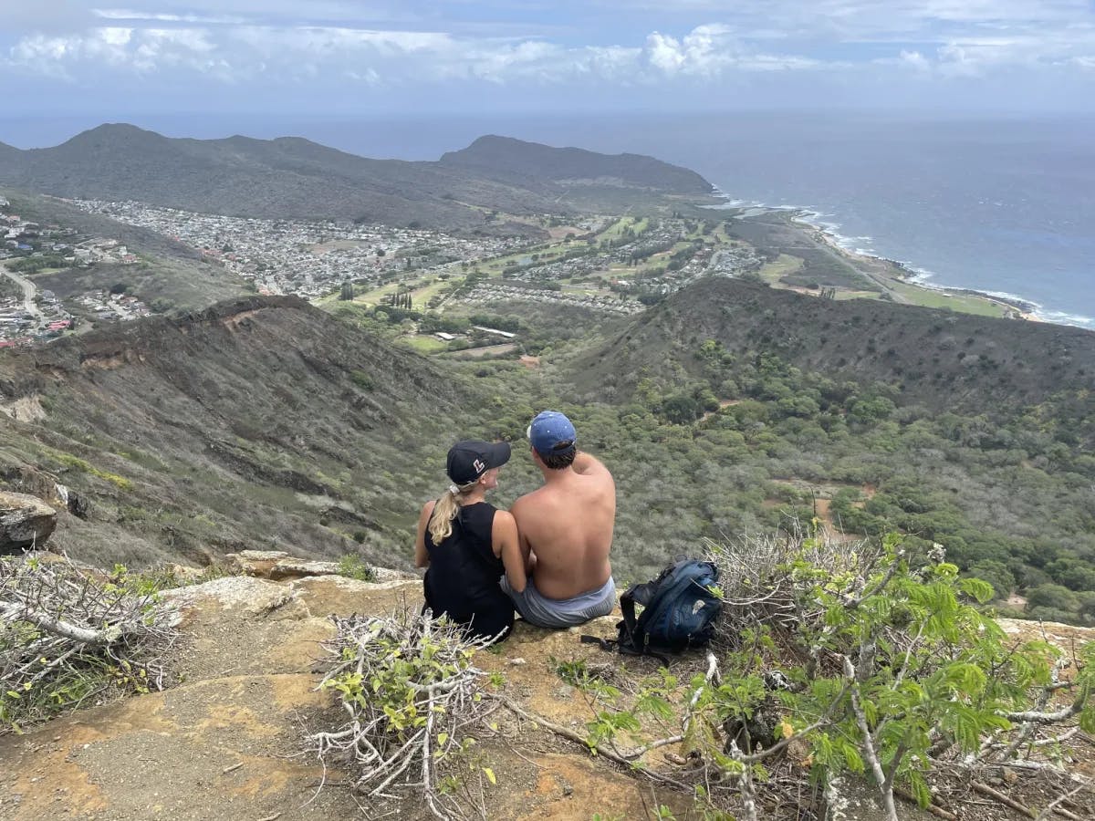 A couple sitting at the edge of a cliff and staring out to a view of mountains and volcanic terrain during a hike.