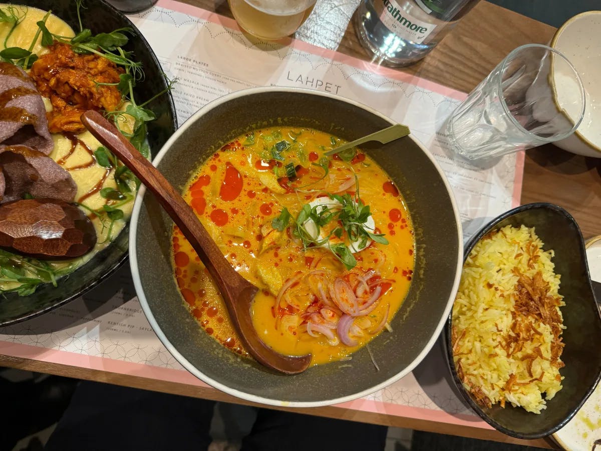 A picture of a table complete with glassware, a menu and three bowls of food that appear to be rice, curry and a meat dish with wooden utensils. 