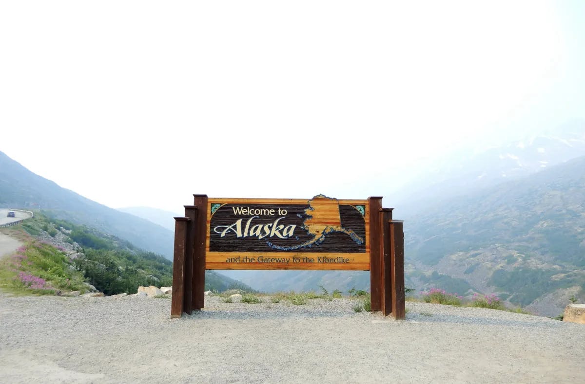 A welcome to Alaska sign on the side of the road with mountains in the background. 