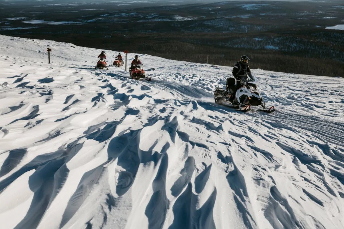 This image depicts four people snowmobiling across a snowy mountain.  