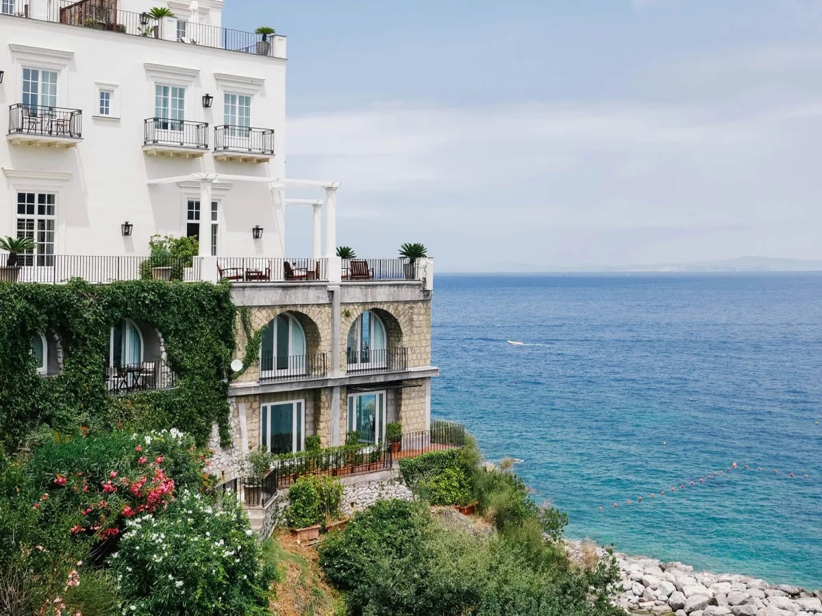 A beautiful white and stone villa looking over the vibrant blue sea and rocky coastline. There are various trees, bushes and plants in the surrounding areas. 