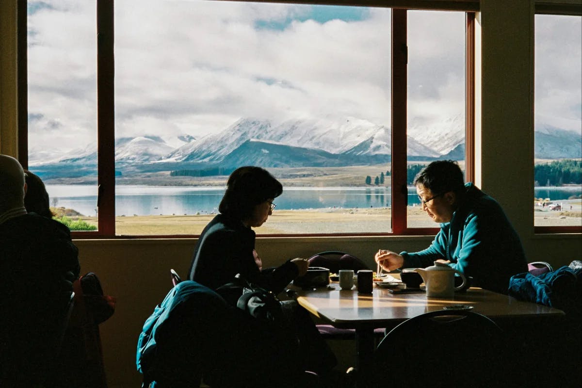 Two individuals are seated at a table in a restaurant with a picturesque view of a lake and mountains through the window.