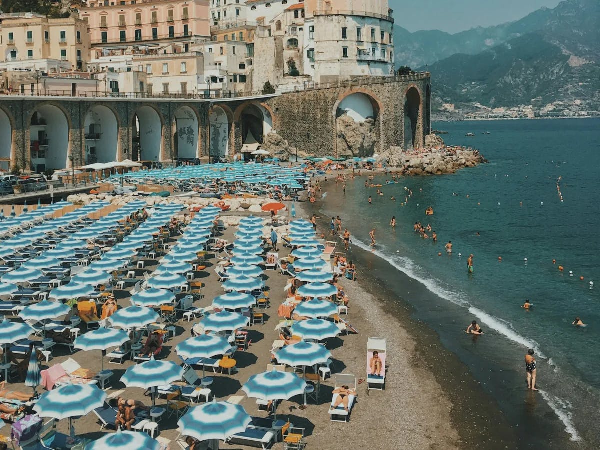 A large grouping of blue and white umbrellas with beach chairs on the pebbly beach with Italian buildings and mountains in the background. 