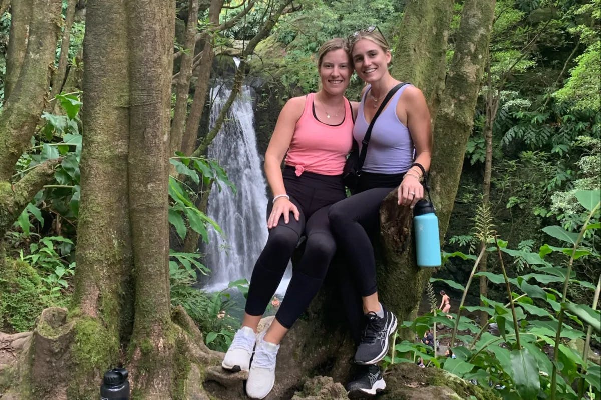 Two girls posing for a photo while leaning on a tree in the jungle. They're wearing a pink and purple tank top, black leggings and sneakers. There is also a waterfall behind them. 