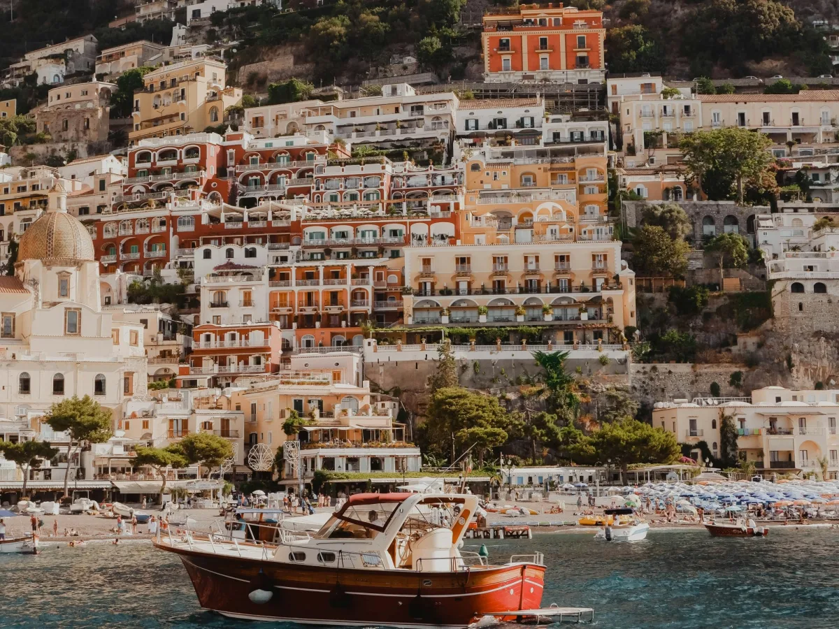 A red and white boat floating on the water with a colorful coastal Italian town in the background. 