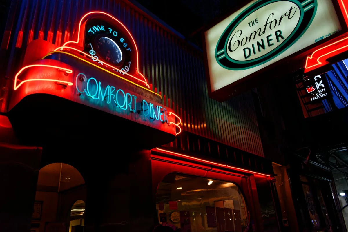The image depicts the illuminated neon signs of “The Comfort Diner” at night, with two prominent signs—one oval and another clock-shaped—above the entrance