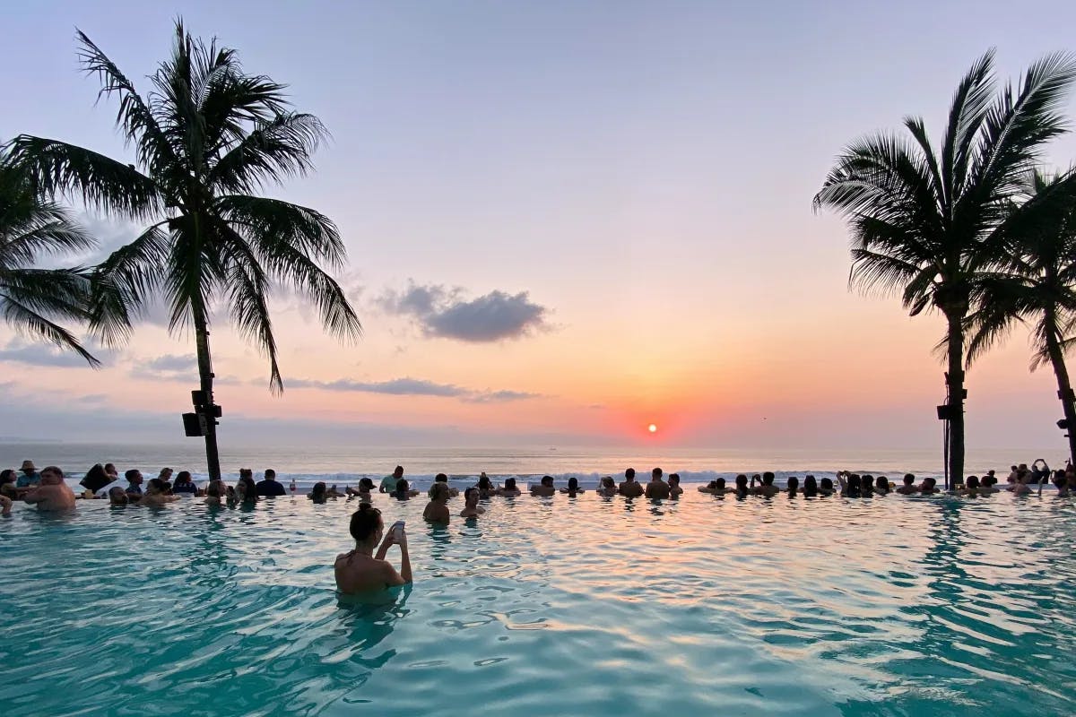 A picture of people swimming in the water near coconut trees in a swimming pool at sunset.