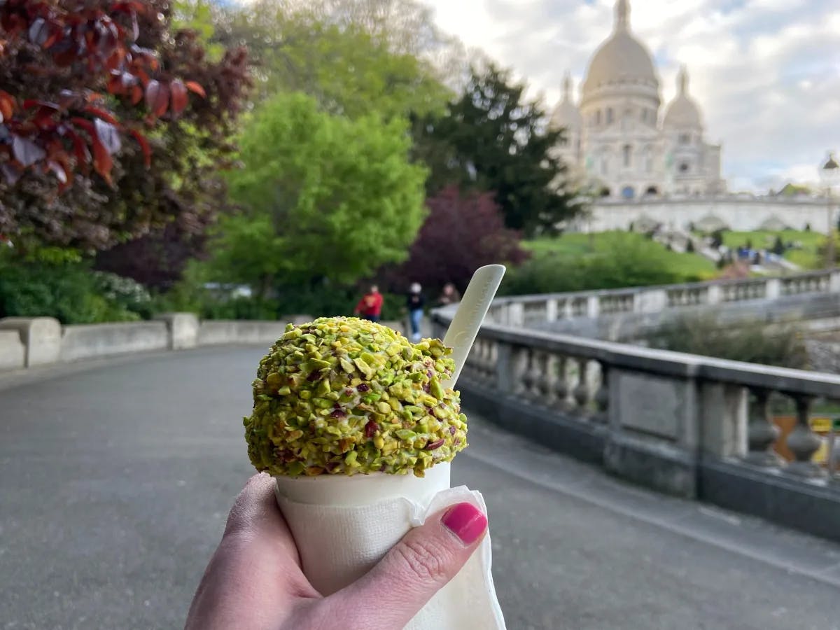 The image showcases a hand holding a pistachio-covered ice cream cone against a blurred backdrop of an urban park and Sacré Coeur Basilica. 
