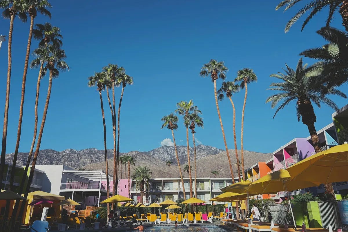 A poolside scene with yellow umbrellas, colorful terraces and tall palm trees in front of a mountain.