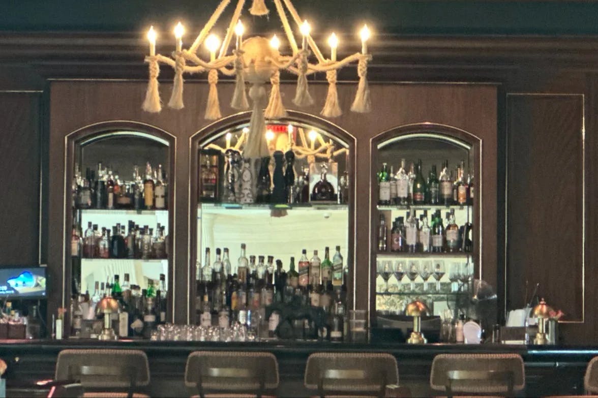 A wooden bar with alcohol stored on the shelves and a gold chandelier above the bar and a piece of artwork. 