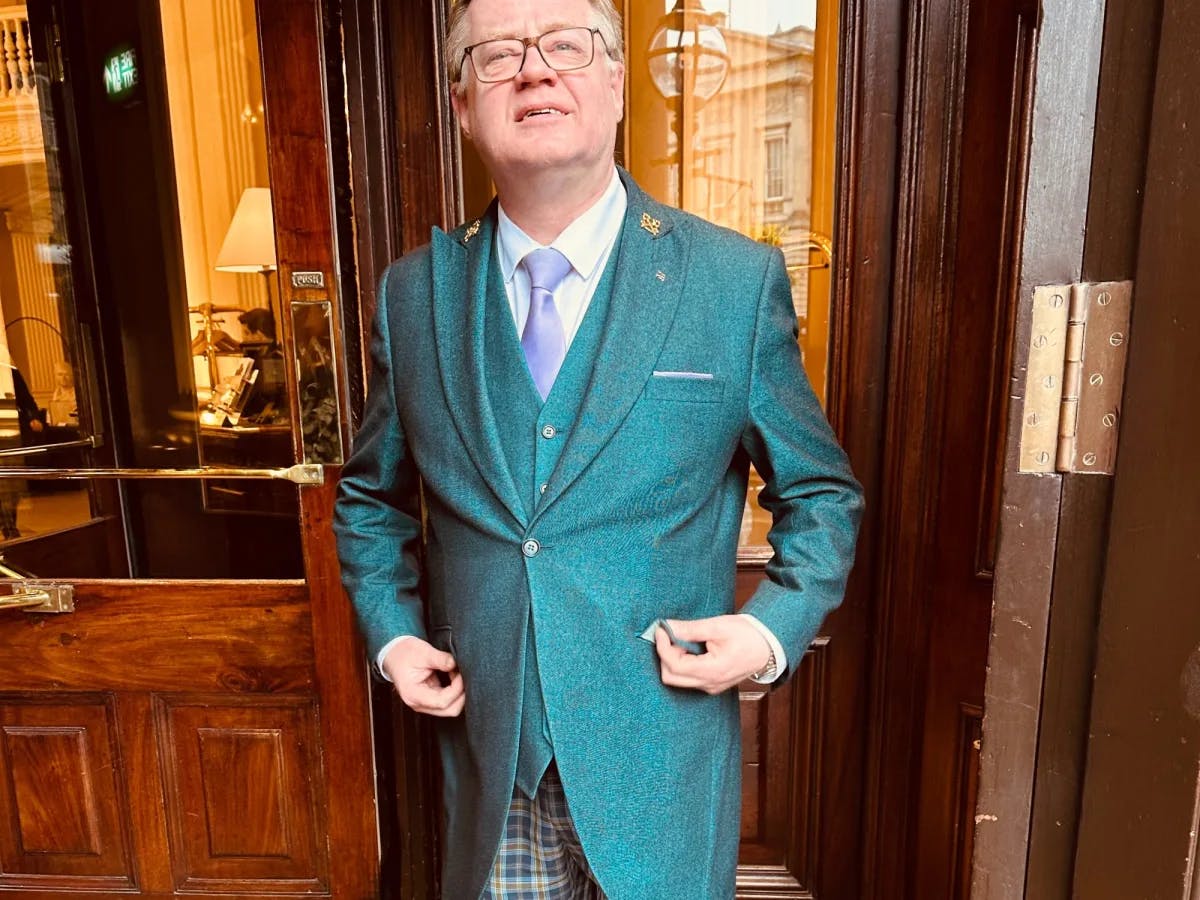 The doorman of a hotel, wearing a green waistcoat, blue tie, checkered pants, and glasses.