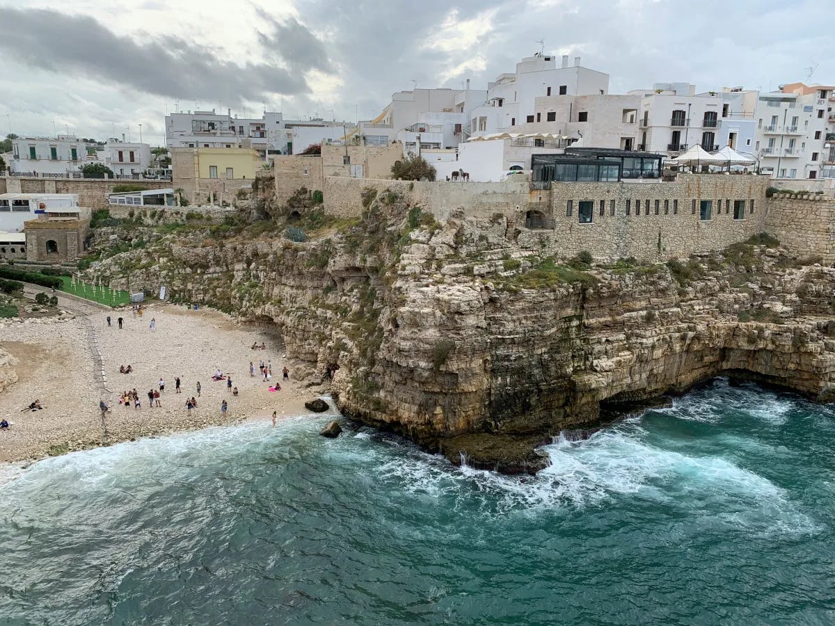 An aerial view of the Polignano a Mare, with a view people swimming, snorkeling and cliff diving amidst the rocky and sandy shoreline and buildings. 