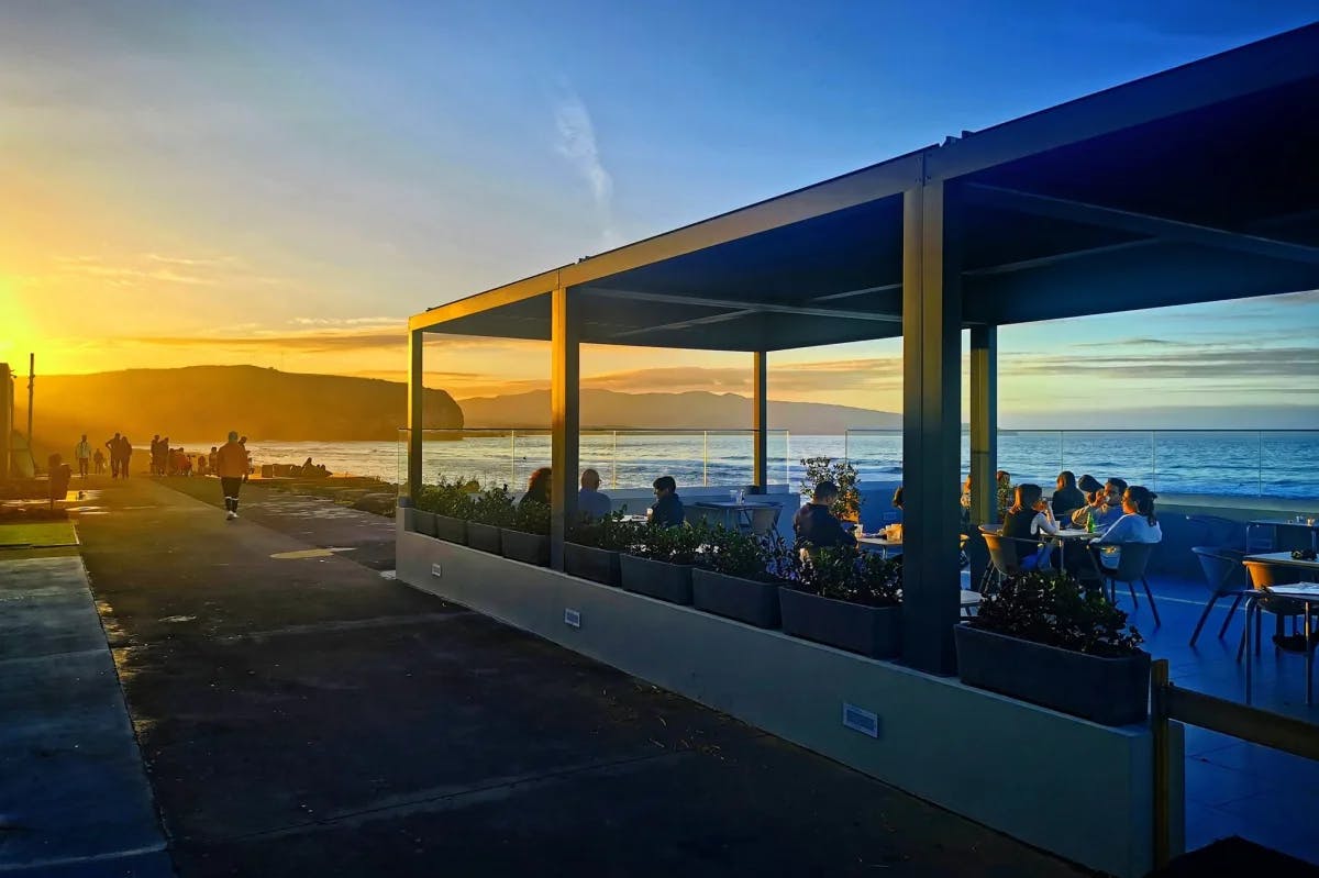 A view of the TukáTulá Beach Bar. There are people dining under a roof to the right with the sea in the background and a golden sunset over the mountains on the left. 