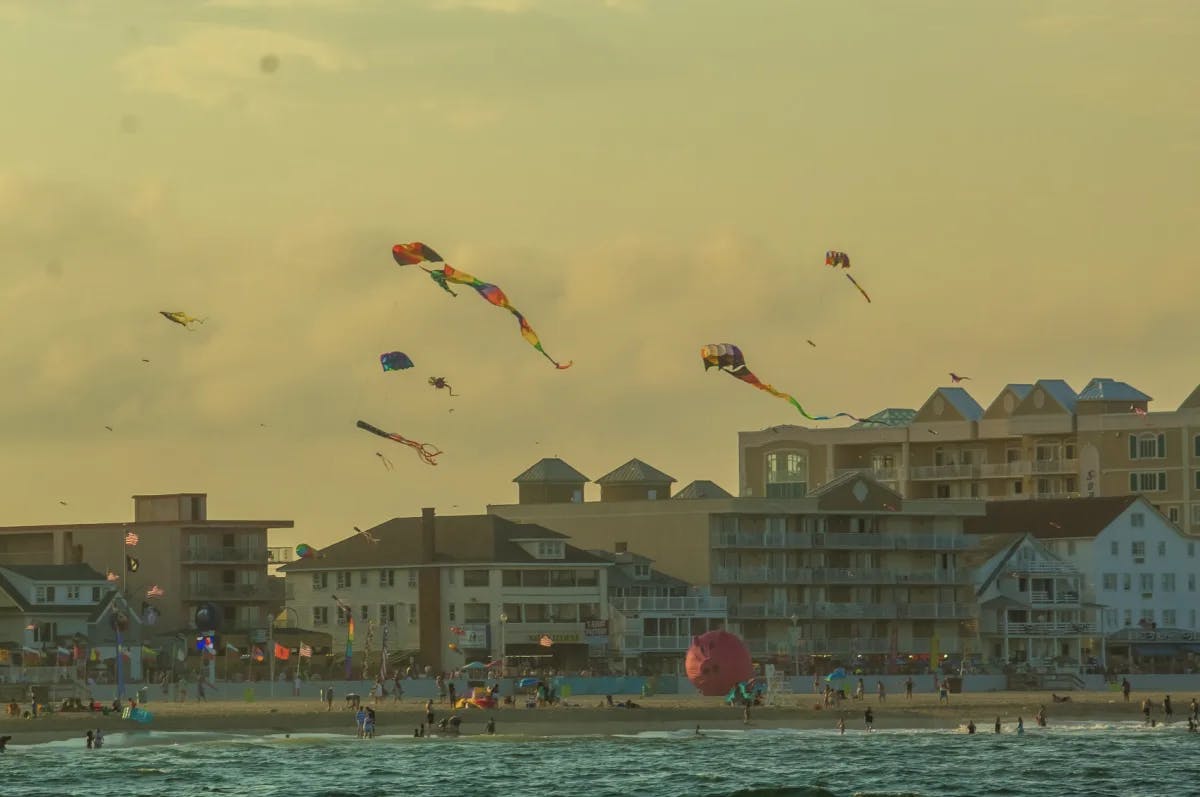 A view of the beach, kites and people walking along the beach with buildings in the background.  