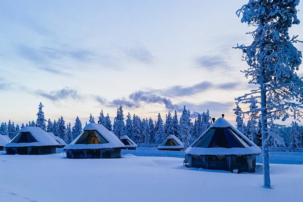 This image depicts multiple snow covered huts with snow covered pine trees in the surrounding area beneath a cloudy sky. 