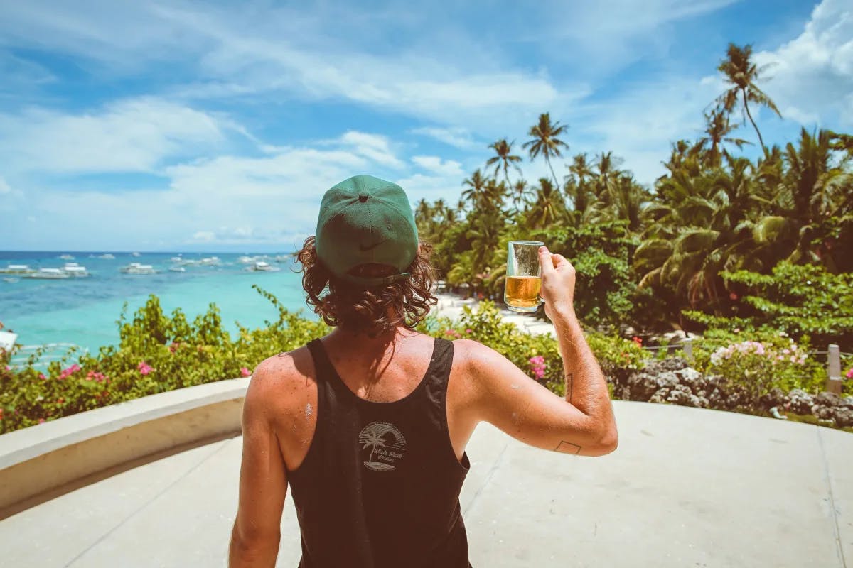 A person in black tank top holding a beer and looking out at the beach.