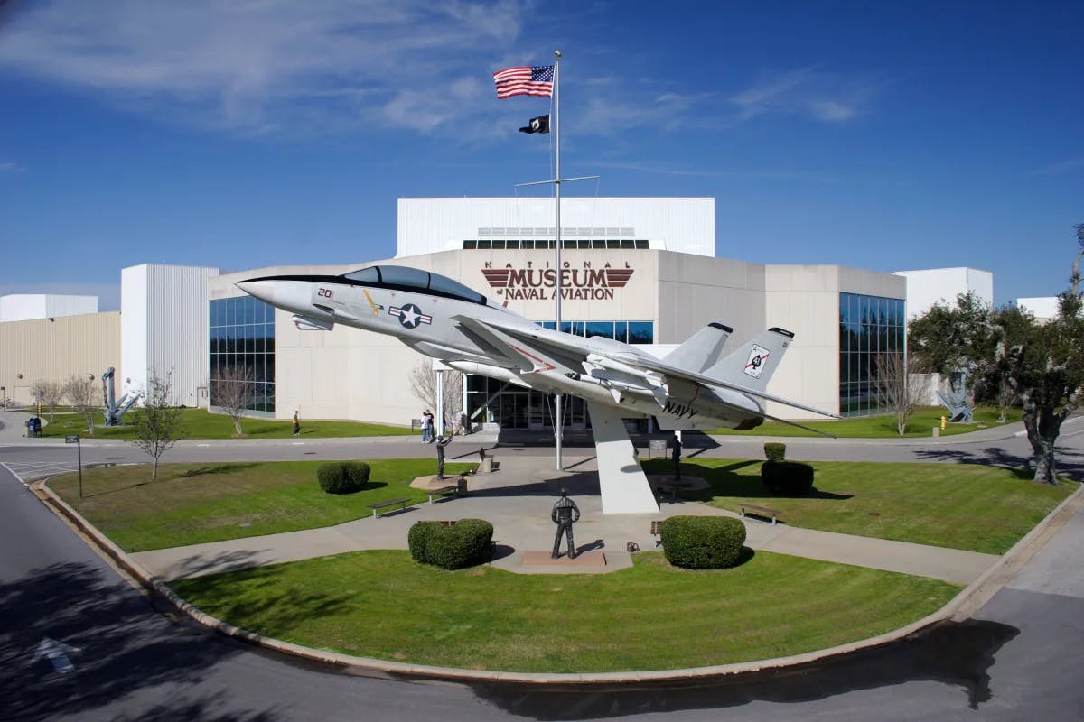 A view of the National Naval Aviation Museum with an airplane in front of a white building with an American flag and grass lawn in the surrounding areas. There is also a street and various sidewalks. 