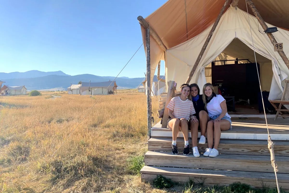 Tori and her friends in casual clothes sitting on steps outside of a glamping tent in a golden field with blue sky overhead.