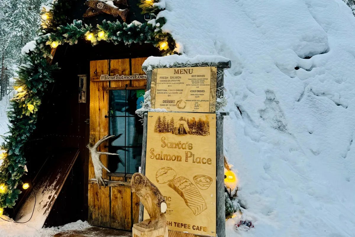 A wooden menu reading "Santa's Salmon Place" outside of a teepee-shaped, snow-covered wooden restaurant with greenery around the trim