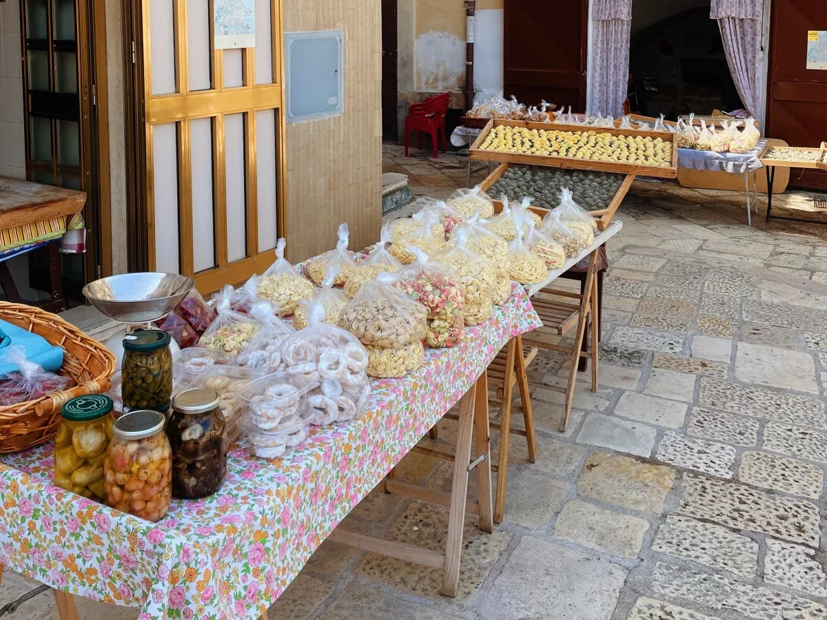 A street view of food stored in bags and jars on top of a wooden tablecloth. There is a wooden door and stone walls in the background. 