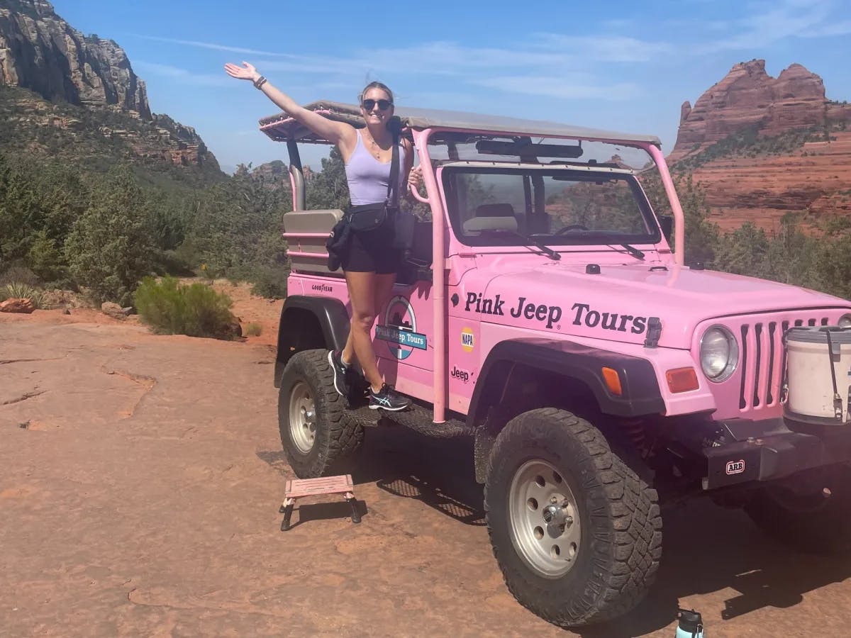 A woman standing on a pink jeep with red rocks in the background.