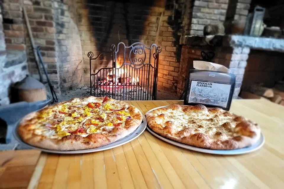 Two pizzas on a platter on top of a wooden table with a metal gate in front of a fireplace in the background. 