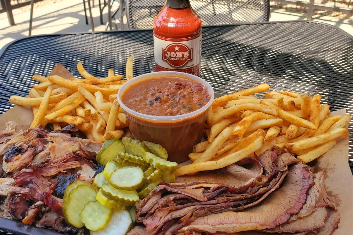 A platter full of meat, french fries, sauce and a jar of hot sauce on a patio table outside. 
