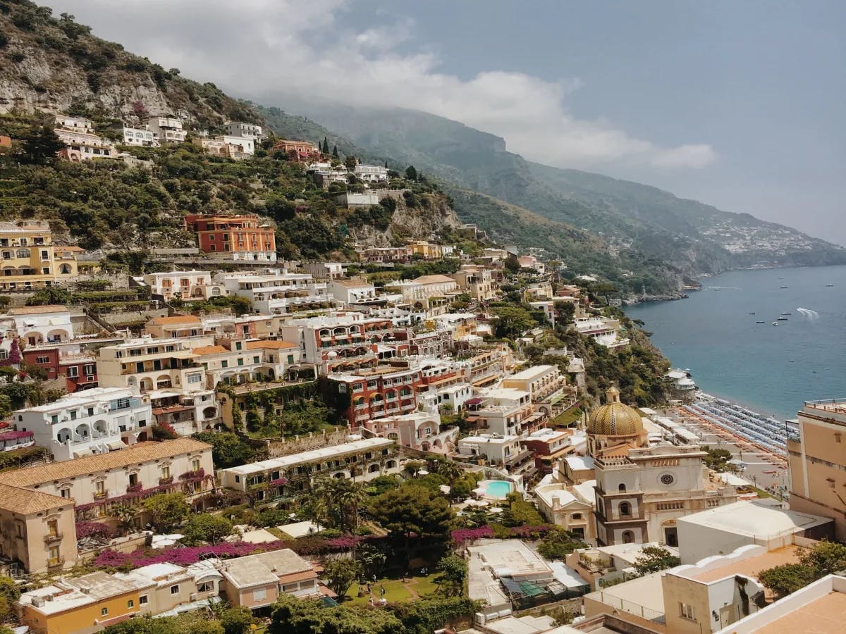 A picture of the coastal town of Positano, with various buildings nestled in the mountainside. There are trees scattered throughout, an umbrella filled beach and the sea on the right side of the image. 