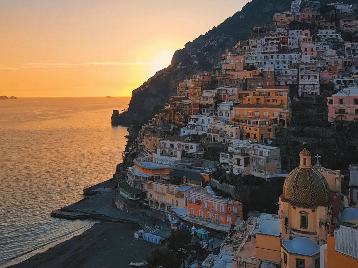 A beautiful view of a golden sun setting over the sea behind a cliff full of a colorful and coastal Italian town. 
