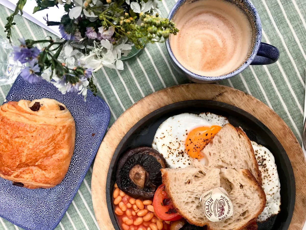 A wooden platter with beans, meat, toast and eggs on top of it. There is a chocolate croissant on a blue dish next to the wooden platter, flowers in the upper left hand corner and a cappuccino towards the right hand corner. 
