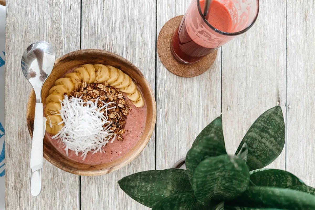 An overhead view of a smoothie bowl, spoon and smoothie glass next to a plant. 