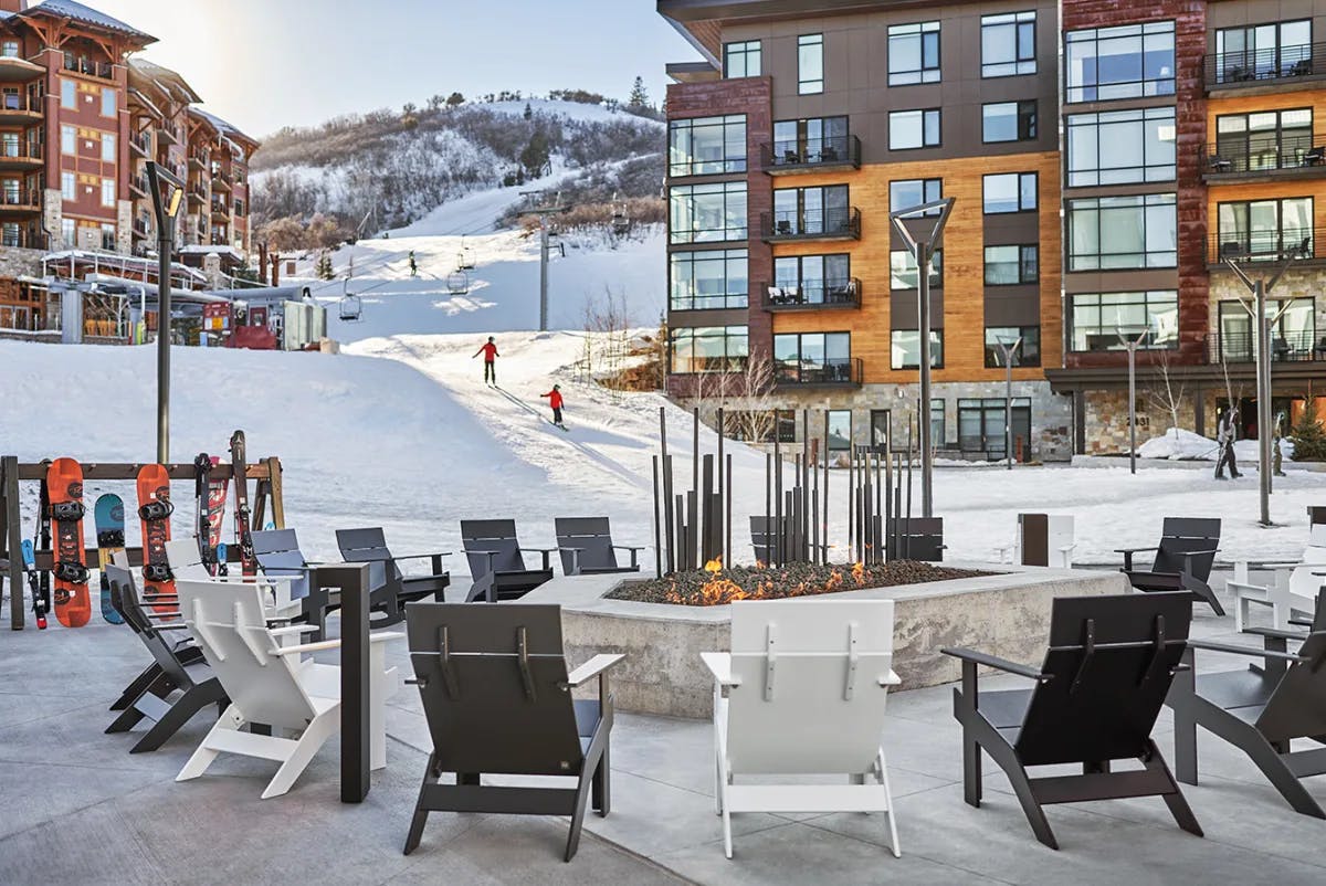 An outdoor seating area around a fire pit with hotel buildings and people skiing in the distance