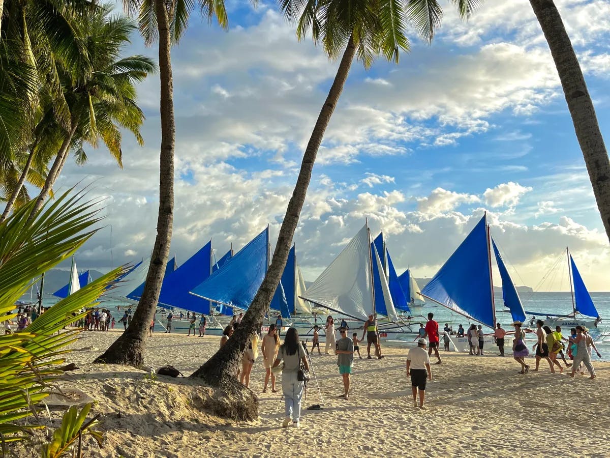 A group of people walking along the beach during the daytime. There are palm trees and blue and white sailboats in the surrounding areas. 